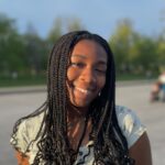 Ameerah Adetoro A young woman with long, braided hair, smiling and wearing a light-colored top with butterfly patterns, standing outdoors with trees in the background.