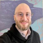 Joshua Dietz E_1_201_a A man with a shaved head, beard, and glasses, smiling and wearing a black sweater over a collared shirt, standing in front of a colorful abstract background.
