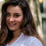 Morella Harris A young woman with long, dark hair, smiling, and wearing a white blouse, standing outdoors with trees in the background.