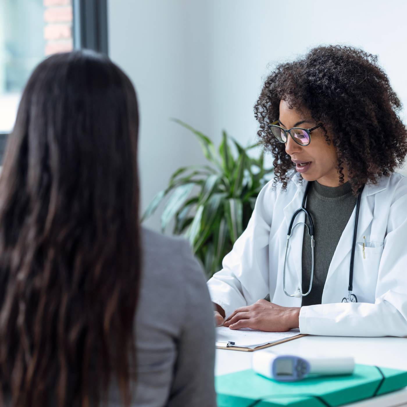 Doctor wearing a white coat and stethoscope speaking with a patient across a desk during a medical consultation.