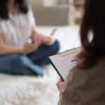 Therapist holding a clipboard and taking notes while listening to a client in a calm, home-like counseling setting.