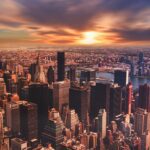 Aerial view of a cityscape at sunset with numerous skyscrapers, including the MetLife Building and the Chrysler Building, and a river running through the city. The sky is filled with dramatic, colorful clouds.