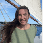 Sarah Lynch A woman with long, brown hair, smiling, and wearing a light green top, standing on a boat with sails, with a body of water and blue sky in the background.