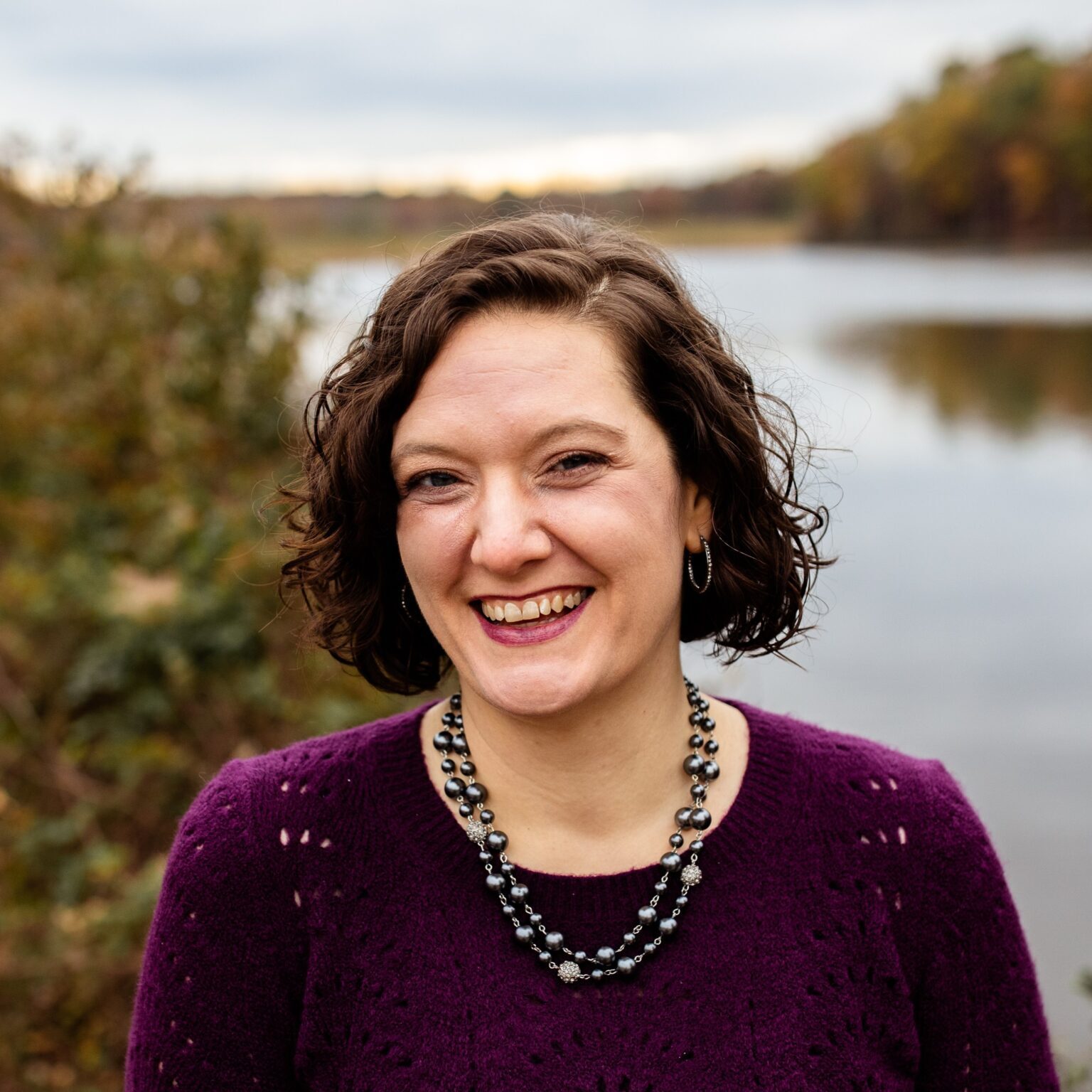 Stephanie Jensen headshot photo A woman with short, curly brown hair smiles broadly at the camera. She is wearing a purple sweater and a layered bead necklace. The background shows a calm river and autumn trees.