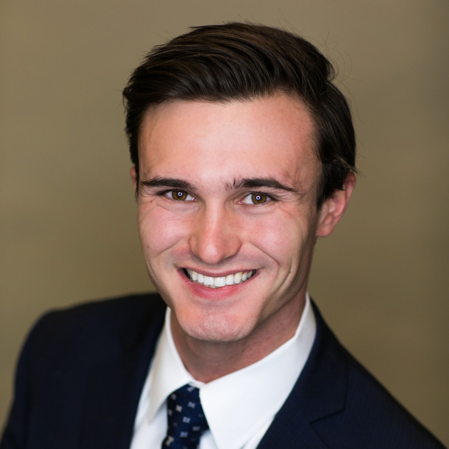 Braeden Kelly Headshot A man with neatly styled brown hair and a big smile looks directly at the camera. He is wearing a dark blue suit and a white shirt with a polka-dotted navy tie.