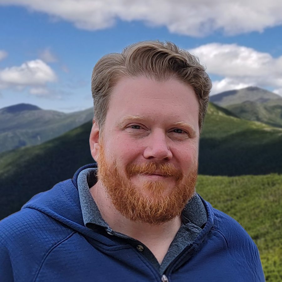 Jason Lutjen photo A man with short, light brown hair and a red beard smiles slightly at the camera. He is wearing a blue jacket, and the background shows green mountains under a partly cloudy sky.