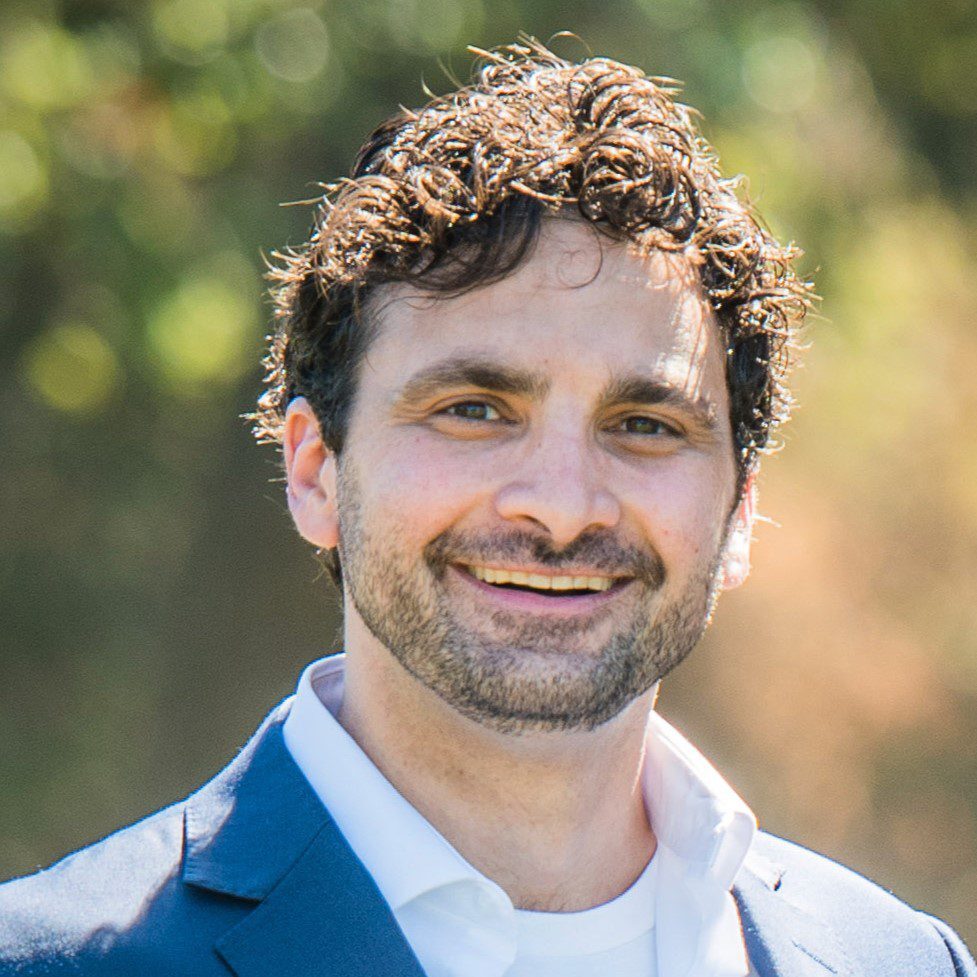 Noah Painter Davis A man with curly brown hair and a short beard smiles at the camera. He is wearing a blue jacket over a white shirt, and the background is blurred greenery.