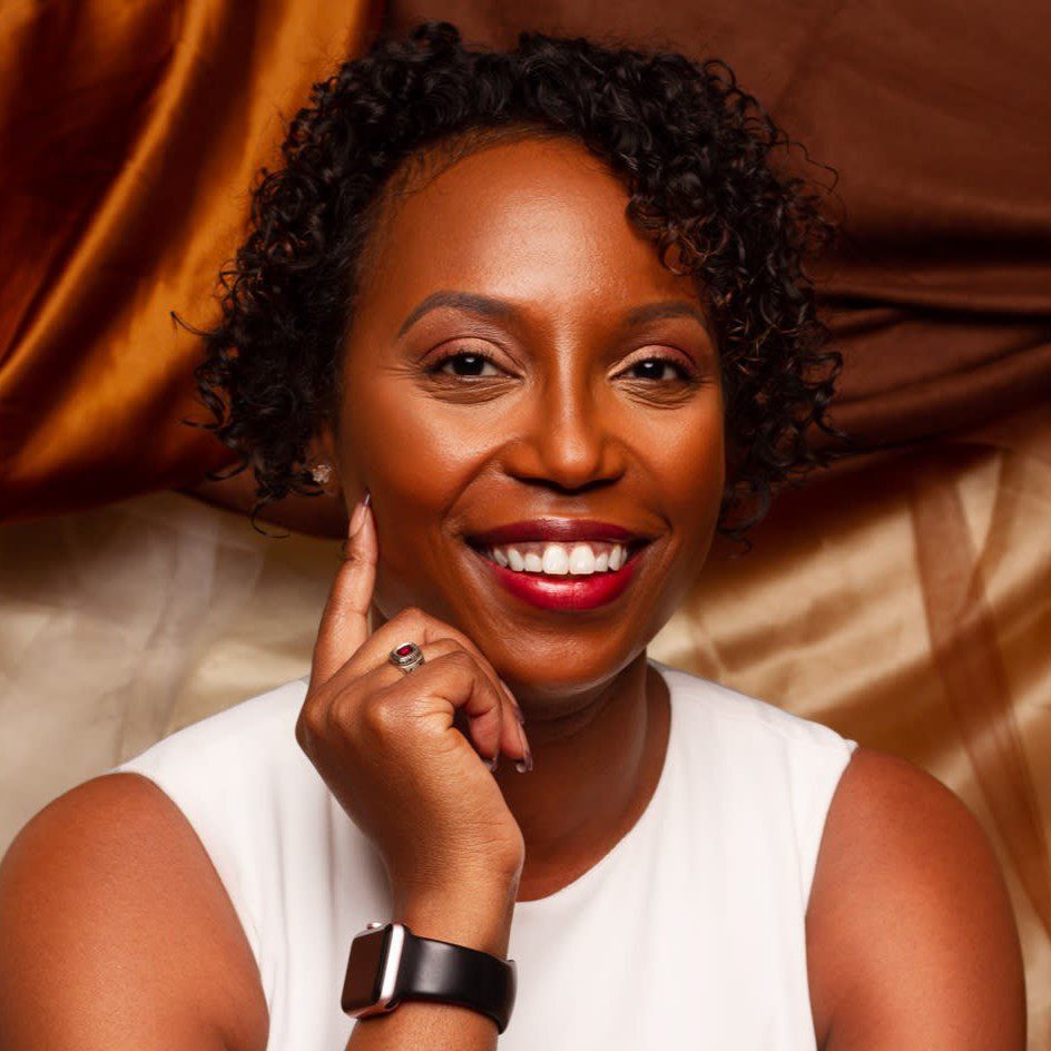 TaLisa Carter headshot A smiling woman with short curly hair wearing a white top and a black smartwatch. She rests her chin on her hand, with a ring visible on her finger. The background features brown and beige draped fabric.