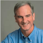 A man with short grey hair smiling and wearing a blue dress shirt, set against a gray background.