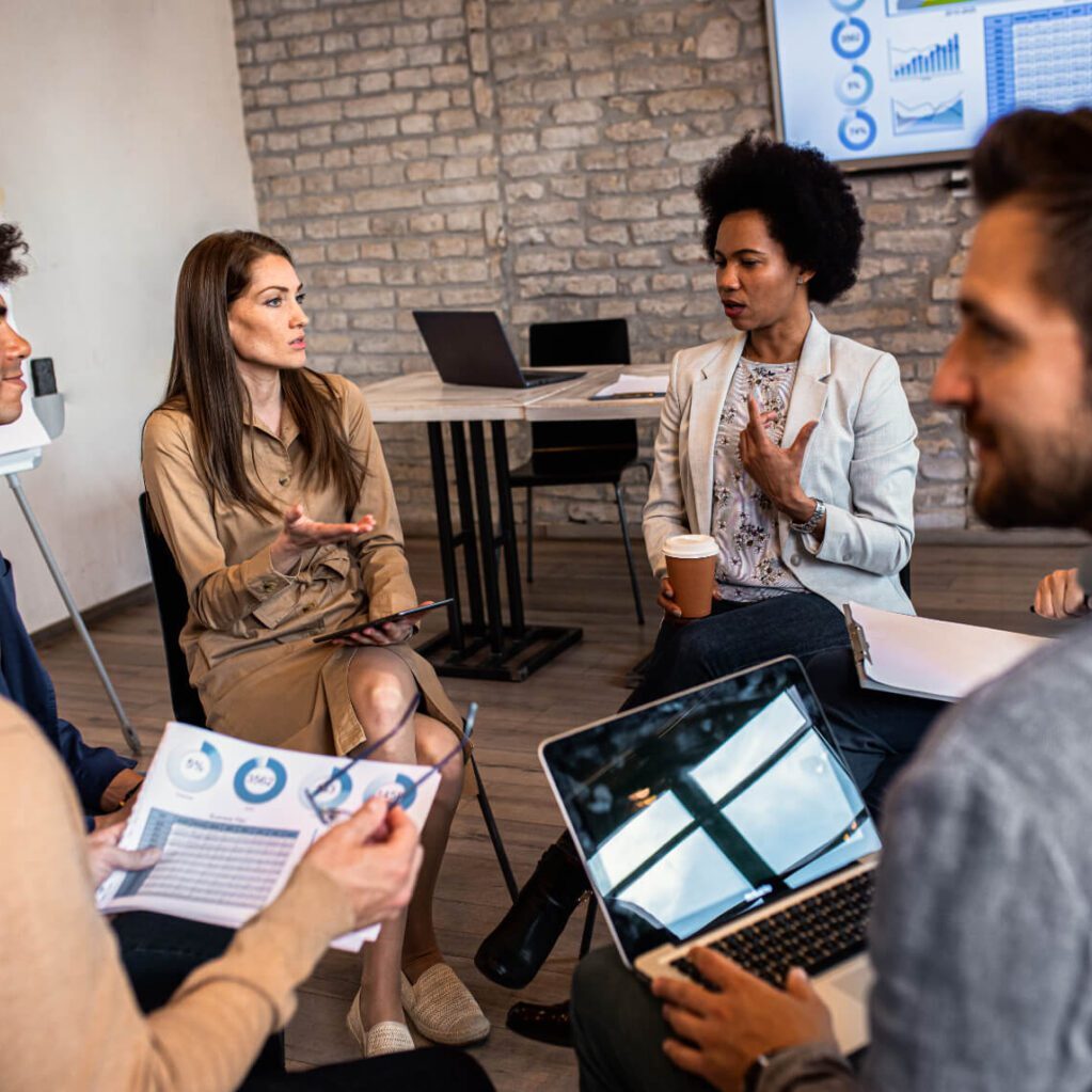 Diverse group of professionals seated in discussion, reviewing charts and laptops.