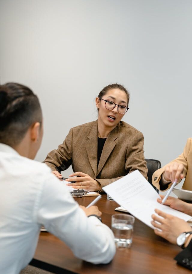 Professional Discussion During Business Meeting A woman in a brown blazer speaks during a meeting, holding documents while seated at a conference table.