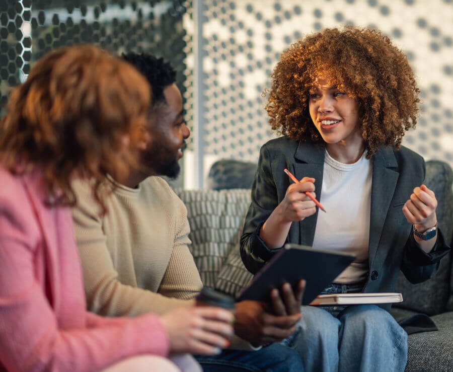 Three people engaged in a discussion while sitting on a couch. The central figure, a woman with curly hair, is animatedly talking and holding a pen and a clipboard. The others, a man and a woman, are listening attentively.