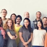 A diverse group of 16 people smiling and standing together against a white background. The group includes individuals of various ages, ethnicities, and styles.