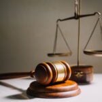 A wooden gavel resting on a sound block in the foreground with a set of brass scales in the background, symbolizing justice and law.