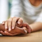 Close-up of two hands on a table, one person’s hand comforting the other by placing their hand on top.