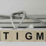 Letter tiles spelling out the word "stigma". A pile of books with a stethoscope draping over them is blurred in the background.
