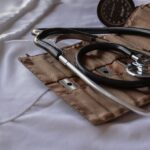A close-up image of medical equipment on a white background. The items include a stethoscope with a shiny metal chestpiece and black tubing, and a brown, weathered leather pouch possibly containing other medical tools.