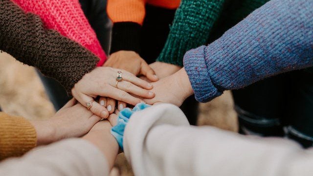 A group of people with hands joined together in a circle, showcasing unity and teamwork, set against a backdrop of autumnal outdoor clothing.